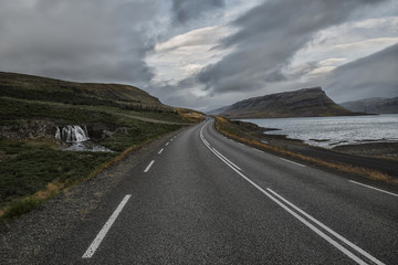 a deserted asphalt road running away into the hills, along the road a waterfall. Iceland. The spirit of travel and adventure.
