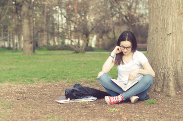 Beautiful young school or college girl with long hair and eye glasses sitting on the ground in the park reading the books and study for exam