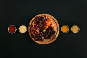 top view of delicious sliced assorted meat on wooden board with various sauces on black