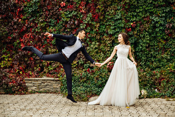 Just married couple in front ol old brick wall with ivy, the bride holds groom's hand and he jumps