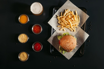 top view of french fries and tasty burger on tray, glass of beer and assorted sauces on black