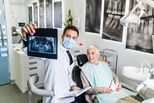 Handsome And Attractive Male Dentist Looking At Dental X-ray Together With His Beautiful Senior Woman Patient.