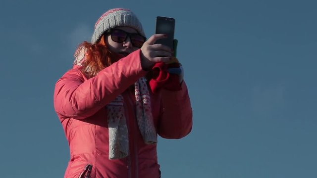 Attractive redhead girl in sunglasses makes selfie on her smartphone. Woman makes photos on mobile phone. Unidentifiable view. Could be anywhere. A girl in bright clothes, blue sky