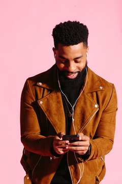 Cool African American man with beard texting, smiling and looking down at his phone, isolated on pink studio background