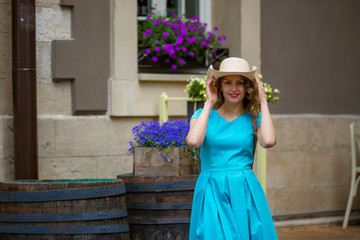 Young woman smiling looking camera, in blue dress, holding hat, outdoor, beautiful city, purple flowers