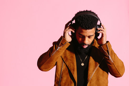Cool African American man with beard holding headphones and looking down, isolated on pink studio background