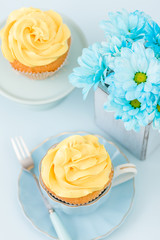 Cupcake with tender yellow cream decoration and bouquet of blue chrysanthemum in retro shabby chic vase on blue pastel background.