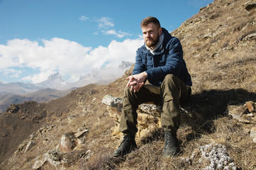 Naklejka premium A portrait of a stylish bearded hipster sitting on a rock against the backdrop of epic rocks and contemplating into the distance thinking about life. The concept of tranquility and solitude with