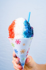 Red, white and blue snow cone in paper cone and blue straw held by female hands.