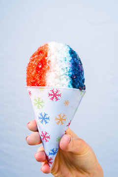 Red, white and blue snow cone in paper cone and blue straw held by female hands.