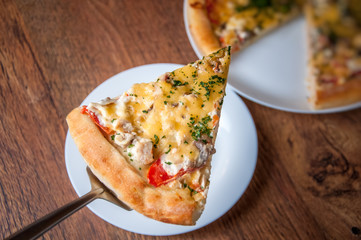 A triangular piece of pizza is taken out of it. the rest of the pizza in blur in the background.Home made pizza on white plate dish on dark oak wood table