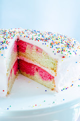 An ombre vanilla and pink birthday cake with slice cut out on white cake stand with multicolored sprinkles and blue background. © Jacob
