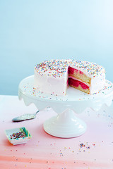 An ombre vanilla pink birthday cake with slice cut out on white cake stand with multicolored sprinkles and pink background on pink paper surface with candles and sprinkles in striped side dish. © Jacob