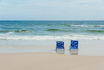 Fototapeta na wymiar Two bright blue beach chairs rest on a sandy beach as foamy blue waves crash towards shore.