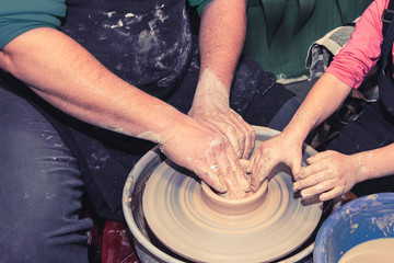 A close-up of a man potter teaches a child how to properly mold a bowl of brown clay on a potter's wheel in a bright workshop, a top view.