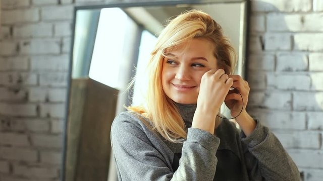 portrait of beautiful young pretty blonde woman taking off her earring and alluring raising moving eyebrows looking camera charming smiling sunny room mirror happy emotional face nice makeup smile