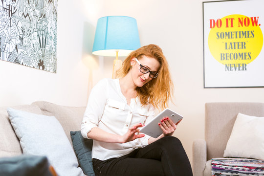 Beautiful young woman with long red hair and glasses for vision uses a white tablet sitting at home in a bright room on the couch with pillows on the floor lamp and pictures on the wall.