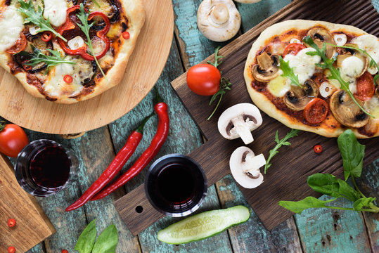 Pizza party flatlay. Rustic pizzas with bell pepper, mushrooms and arugula served with raw chili pepper, champignons, tomatoes and red wine on shabby blue background