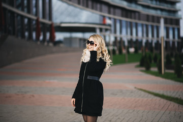 Fototapeta premium Beautiful young girl with blond wavy hair in a black coat against the backdrop of modern buildings