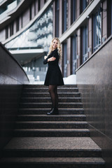Beautiful young girl with blond wavy hair in black dress goes against the background of modern building