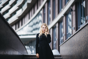 Obraz premium Portrait of a Beautiful young girl with blond wavy hair in black dress on background of modern building