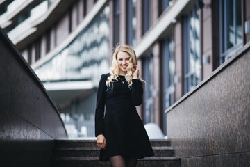 Fototapeta premium Portrait of a Beautiful young girl with blond wavy hair in black dress on background of modern building