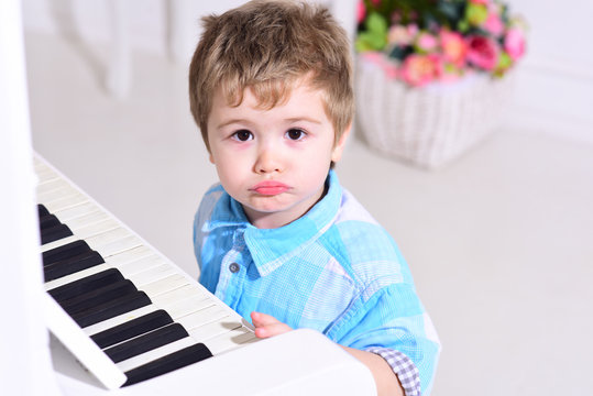 Child sit near piano keyboard, white background. Boy cute and adorable puts finger on keyboard of piano. Kid spend leisure near musical instrument. Elite childhood concept.