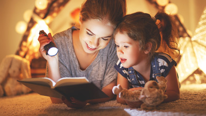 mother and child daughter reading a book and a flashlight before going to bed