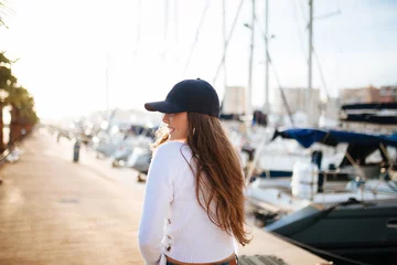 Fotobehang Attractive young woman posing with a cap in marina in the sunset © ManuPadilla