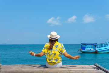 Fototapeta na wymiar back view of beautiful asian female in fashion yellow shirt sitting on the edge of pier looking forward to see amazing blue sky with a little cloud, fishing boat and seascape in a sunny day