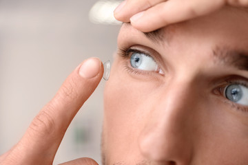 Young man putting contact lens in his eye on light background