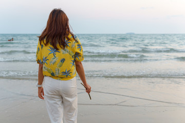 back view of beautiful asian female in fashion yellow shirt walking on the beach looking forward to see amazing blue sky with a little cloud and seascape  in the evening when sunset.