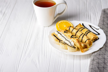 Pancakes with banana, whipped cream decorated with chocolate syrup on white wooden background and cup of tea.