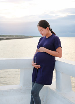 Asian pregnant woman embracing her belly on the seashore at sunset.