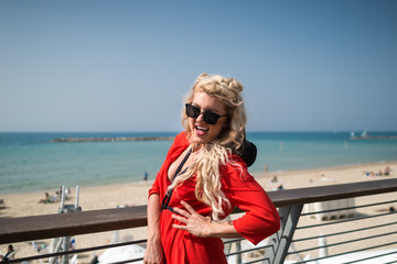 Young smiling woman in bright red dress and sunglass stands on on the city promenade. Trendy blonde girl having fun on the background of beach with relaxing people and sea