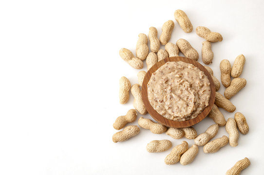 American Breakfast, Sweet Food And Snack High In Calories Concept With A Bowl Of Crunchy Peanut Butter Surrounded By Peanuts Still In Shell, Isolated On White Background With Copy Space