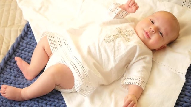 A cute kid lies on a bed in a shirt for baptism, moves his arms and legs, studies the world around, smiles, looks at the camera. HD, 1920x1080. slow motion.