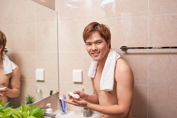 Young handsome asian man applying cream onto face in bathroom