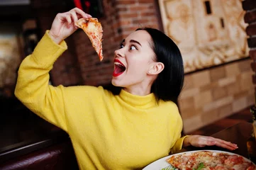 Crédence de cuisine en verre imprimé Pizzeria Drôle de fille brune en pull jaune, manger de la pizza au restaurant.
