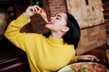 Foto auf Acrylglas Pizzeria Lustiges Brunettemädchen im gelben Pullover, der Pizza im Restaurant isst.