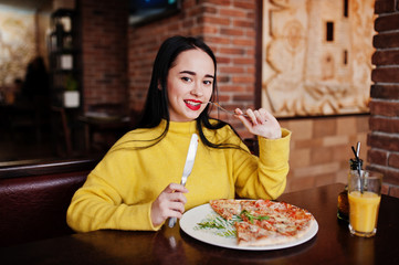 Funny brunette girl in yellow sweater eating pizza at restaurant.