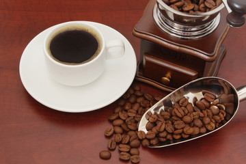 White cup of coffee, manual coffee grinder and roasted beans, metallic shovel on wooden