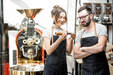 Couple of baristas in uniform checking the quality of roasted coffee beans standing near the roaster machine