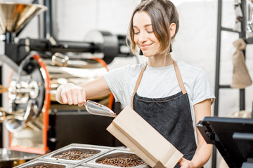 Young woman seller in uniform filling bag with coffee beans working in the coffee store