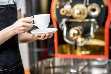 Waiter in uniform holding a coffee cup with roaster machine on the background. Close-up view on the cup