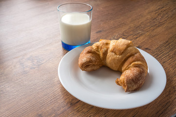 curved croissant on a white plate on a dark oak wooden table and glass of milk