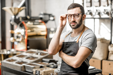 Portrait of a handome bearded barista in uniform standing in the coffee shop with coffee roaster machine on the bckground