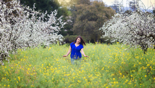 Waist up portrait of young woman in bright blue top in blossoming orchard and yellow mustard field, smiling, looking straight to the camera