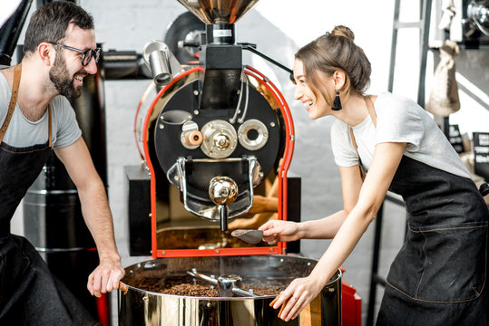 Couple of two baristas checking the quality of the coffee beans standing with scoop near the roaster machine at the roastery
