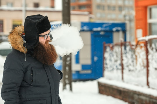 Vape bearded man in real life. Portrait of young guy with large beard in glasses and a black cap vaping an electronic cigarette in the winter.